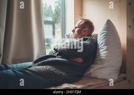Portrait of thinking sad Middle-aged man in eyeglasses dressed open cardigan lying on cozy bed next to window looking on street through raindrop glass Stock Photo