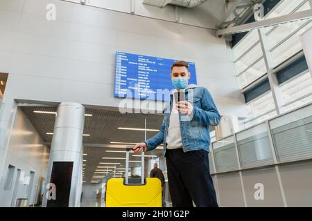 Man in medical face mask using smartphone in airport. Urban traveler wearing casual holds phone during covid lockdown. Man text message holding Stock Photo