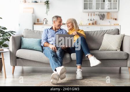 Mature Couple Using Laptop Computer Together Browsing Internet Smiling To Each Other Sitting On Sofa At Home. Senior People And Online Technologies, M Stock Photo