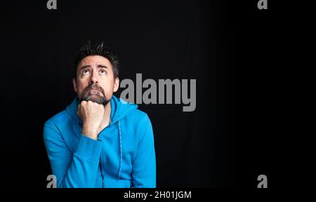 A young man in a blue sweatshirt, dark and bearded, looking up with his fist resting on his chin in a dreamy or pensive attitude. Black background. Co Stock Photo