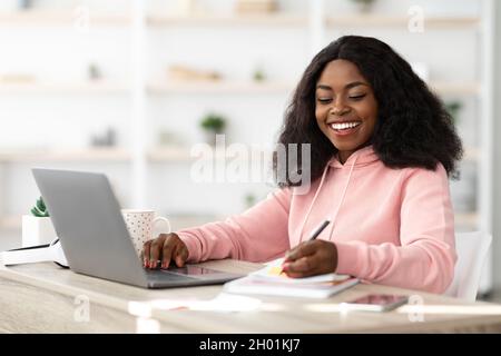 Smart attractive young black woman attending online class from home, sitting at workdesk, using laptop and taking notes, copy space. E-education, trai Stock Photo