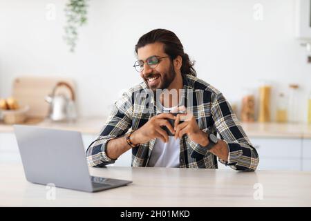 Cheerful indian guy with glasses looking for job online, sitting at table, using laptop, drinking coffee, kitchen interior, copy space. Happy arab man Stock Photo