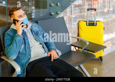 Portrait of attractive young man talking on smart phone in airport lounge with suitcase before traveling by plane. Caucasian man in Protective face Stock Photo