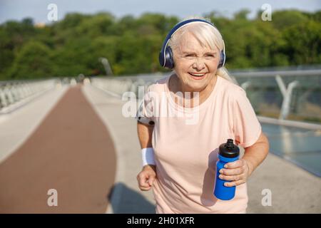 Positive senior lady with headphones runs along bridge in summer Stock Photo