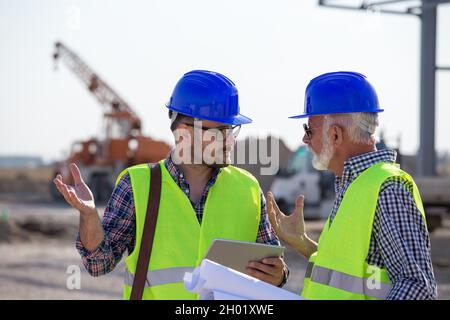 Two engineers with tablet and blueprints talking at building site with metal construction in background Stock Photo