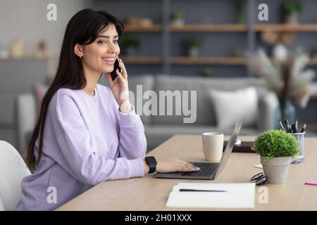 Smiling female employee working on computer in home office and talking on smart phone, happy woman worker using laptop, laughing speaking with friend Stock Photo