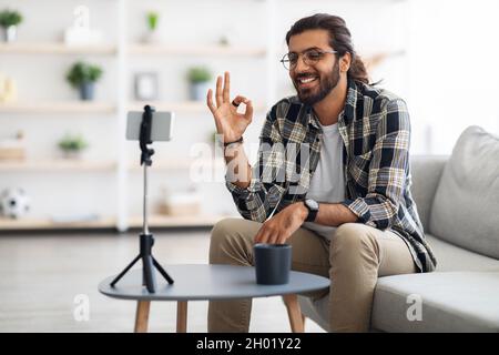 Cheerful indian guy blogger streaming from home, sitting on sofa, looking at mobile phone camera, talking, gesturing, smiling. Middle-eastern vlogger Stock Photo