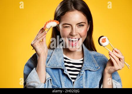Happy smiling girl eating sushi rolls with chopsticks, holding sashimi and winking at camera, order in japanese restaurant, yellow background Stock Photo