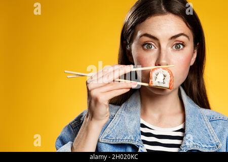 Happy woman orders japanese traditional food makizushi, hold sushi roll with chopsticks and smiling. Girl student eating tasty sushi, standing over Stock Photo
