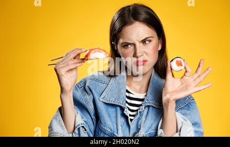 Picky girl looking at sashimi with disgust, dont like raw fish rolls in japanese restaurant, standing displeased against yellow background Stock Photo