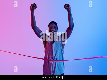 Excited black sportsman running, crossing finish line, raising his arms up in triumph, celebrating victory in neon lighting. Emotional African America Stock Photo