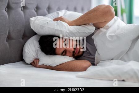 Upset arab man closing his ears with pillow, hiding from too loud sound or noise early in morning, lying in bed. Guy feeling uncomfortable, trying to Stock Photo