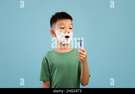 Shocked little asian boy with shaving foam on face looking at razor blade, emotional korean kid wants to be like dad, pretending to be adult man while Stock Photo