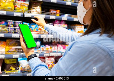 Cropped photo of a young woman with face mask using mobile phone and buying groceries in the supermarket during virus pandemic. Stock Photo