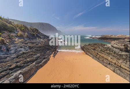 The view of fine sandy beach with rock outcrops and misty sea at Storms River Mouth in Tsitsikamma National Park off the Garden Route, South Africa. Stock Photo