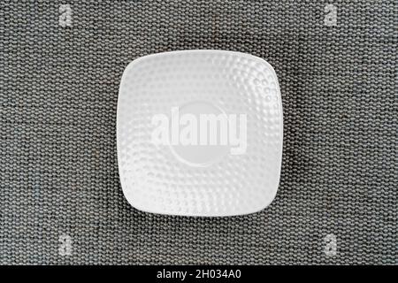 Ceramic white square saucer on a gray rough jute burlap close up. Copy space. Abstract rustic crockery background Stock Photo