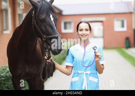 Portrait of young smiling female veterinarian holding steepler while standing next to horse Stock Photo