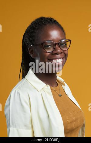 Vertical portrait of young African-American woman wearing glasses and smiling happily at camera while posing against yellow background in studio Stock Photo