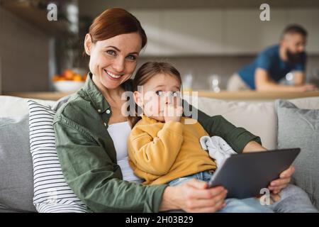 Mother with small daughter using tablet indoors at home, looking at camera, everyday life and home office with child concept. Stock Photo