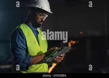 Portrait of happy young industrial man with protective wear using tablet indoors in metal workshop at night. Stock Photo