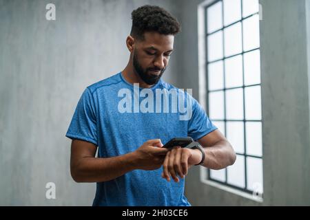 Happy young African American sportsman standing indoors at gym, using smartphone and smartwatch. Stock Photo