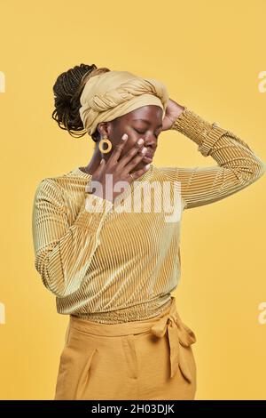 Vertical portrait of trendy African-American woman wearing head wrap and ethnic jewelry while dancing against yellow background in studio Stock Photo