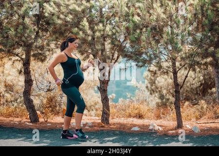 Cute Happy Pregnant Woman on Workout in the Park in Sunny Day. Enjoying Sportive Walk Among Beautiful Fresh Trees. Active Healthy Pregnancy. Stock Photo