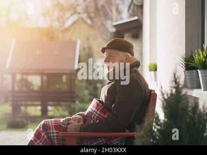 Senior man sitting on bench covered with blanket on porch and enjoying fresh air on cold winter day Stock Photo