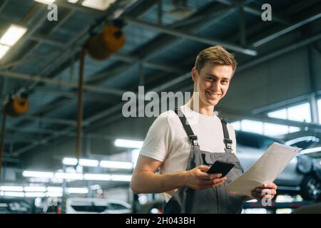 Side view of smiling handsome young mechanic male wearing uniform holding clipboard and using texting mobile phone Stock Photo
