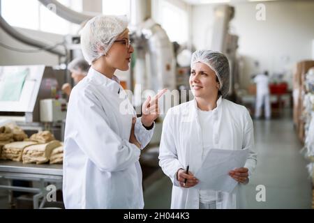 Two female workers discussing while standing in food factory. One of them holding paperwork and pen while other one speaking. Stock Photo