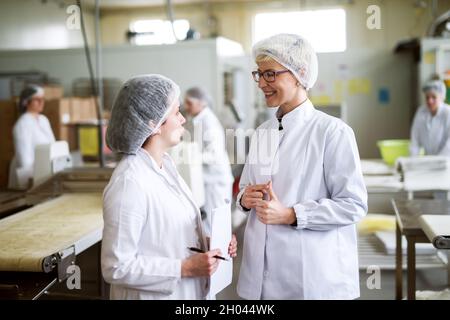 Two female workers discussing while standing in food factory. One of them holding paperwork and pen while other one speaking. Stock Photo