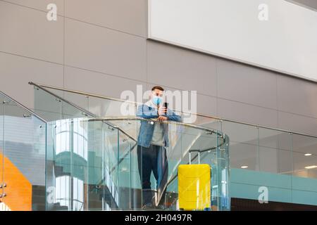 Male traveler Use mobile phone at airport. Man in protective face mask With luggage Use smartphone In airport lounge balcony with outdoor advertising Stock Photo