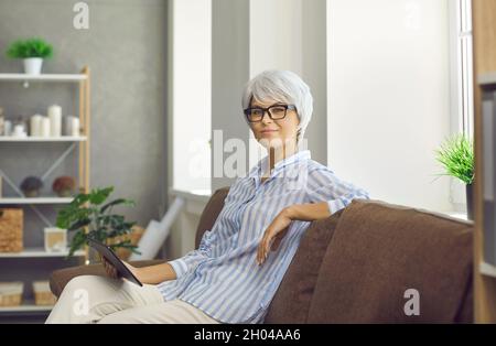 Mature woman with glasses sitting on sofa working or relaxing with digital tablet in hands. Stock Photo