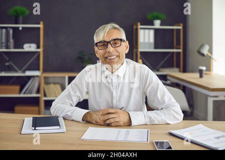 Portrait of happy senior businessman sitting at office table and smiling at camera Stock Photo