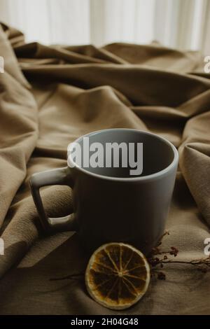 Empty grey coffee cup on beige background isolated while next to it there is a dried plant with a slice of orange. Close-up image. Stock Photo