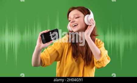 Cheerful redhead teen female in headphones singing song while listening music with audio player app on smartphone, positive young woman, standing over Stock Photo