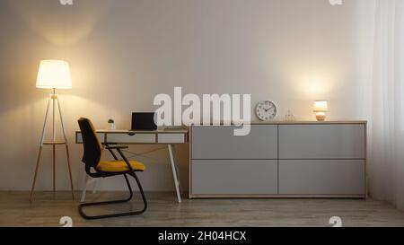 Comfortable armchair, table with laptop with empty screen, furniture with clock, glowing lamps on gray wall background in evening in office or living Stock Photo