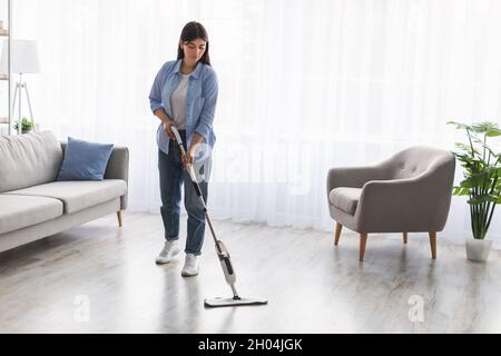 Housewife. Casual woman washing hardwood laminate flooring in living room using water spray mop pad and refillable bottle with cleaning agent. Blurred Stock Photo