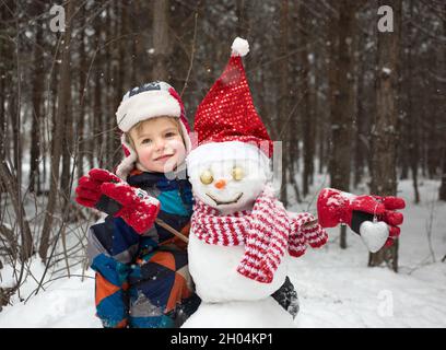 child boy in warm winter overalls and snowman wearing Santa hat, striped scarf and red gloves. Christmas festive season in a snowy forest. Winter walk Stock Photo