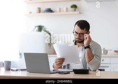 Busy mature caucasian guy with beard in glasses works with documents behind laptop in minimalist kitchen interior. Home bookkeeping, finance, paying m Stock Photo
