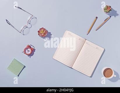 Office or business workspace with open notebook and reading glasses on a blue background with flower, pen, alarm clock, memo pad and mug of coffee in Stock Photo