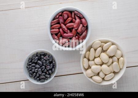 Three kinds of organic red, white, black dried beans in ceramic saucers, close-up, on a white wooden table, top view. Stock Photo