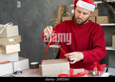 Preparing gifts for Christmas and New Years a man in a Santa hat works in a warehouse preparing an order for goods in boxes for delivery. Sale Stock Photo