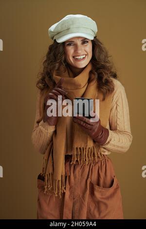 Hello september. smiling modern woman in sweater with leather gloves sending text message using smartphone on bronze background. Stock Photo