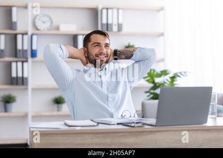 Satisfied emotional middle aged caucasian guy with beard with hands behind head resting from remote work at laptop in office or home. Freelancing, bus Stock Photo