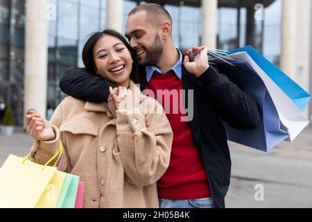 Portrait of happy multiracial couple with shopper bags smiling and hugging near supermarket. Cheerful diverse family going shopping, posing together n Stock Photo