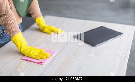 Cleaning Workplace Concept. Closeup of unrecognizable muslim woman wearing protecitve yellow rubber gloves polishing table with laptop using microfibe Stock Photo