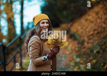 Hello autumn. happy elegant 40 years old woman in brown coat and yellow hat with autumn yellow leaves walking tour outdoors on the city park in autumn Stock Photo