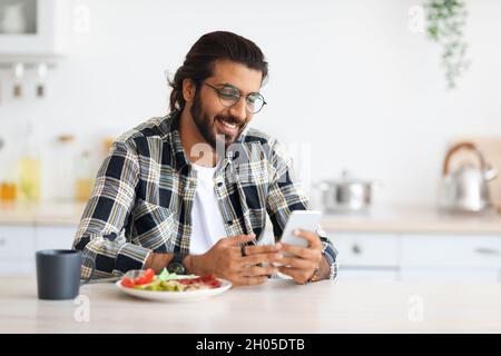 Cheerful bearded long-haired middle-eastern guy with glasses enjoying lunch at home, sitting at kitchen table alone, using mobile phone and smiling, c Stock Photo
