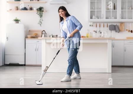 Full body length of cheerful casual woman washing hardwood laminate flooring in kitchen using water spray mop pad and refillable bottle with cleaning Stock Photo